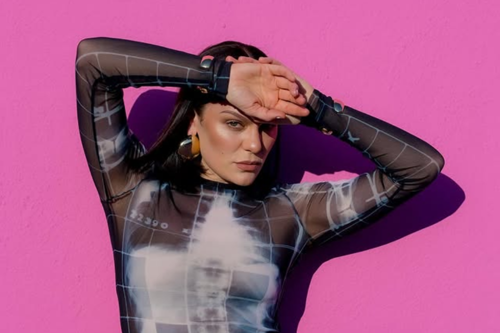Jessie J está com uma mão na frente da testa e outro atrás da cabeça, na frente de um fundo rosa choque