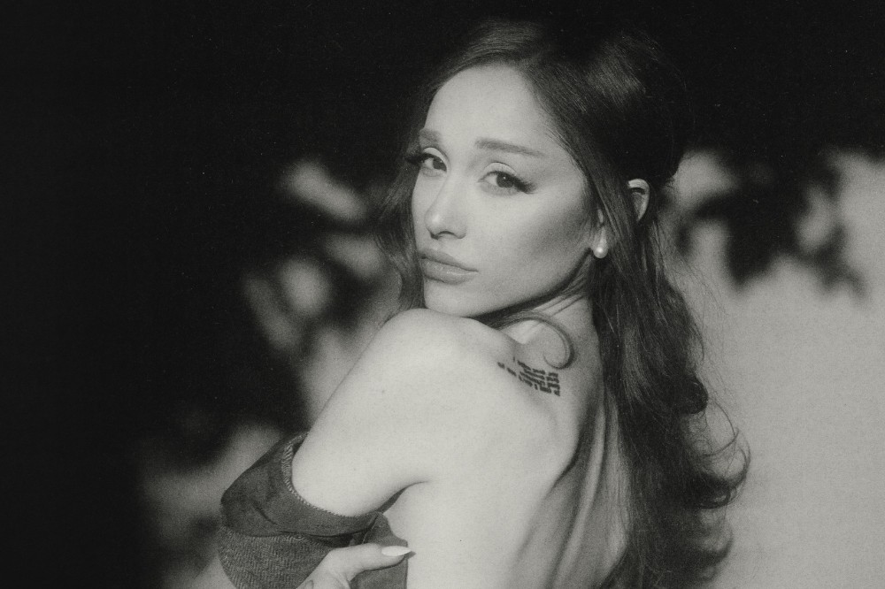 Cantora Ariana Grande em preto e branco, olhando por cima do ombro esquerdo, com cabelo solto e um brinco de pérola. Ela tem uma tatuagem no ombro e veste uma blusa que revela parte das costas